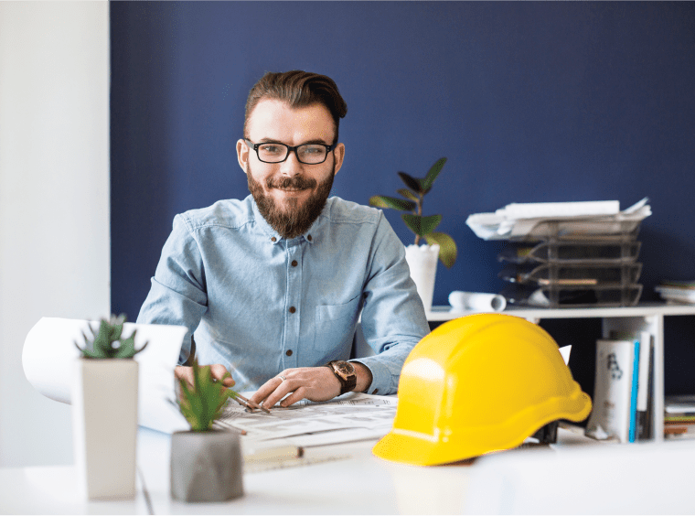 Smiling architect at desk with building plans and yellow helmet.
