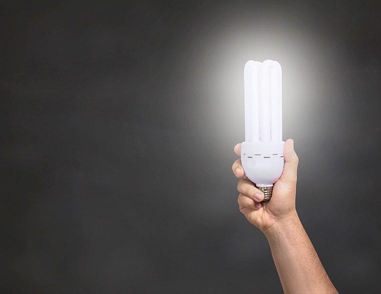 Hand holding a glowing compact fluorescent light bulb against a dark background.