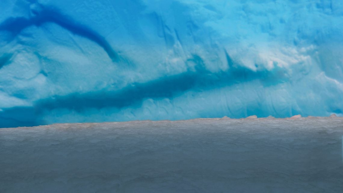 Close-up of a glacier's icy blue wall meeting a snowy ground.
