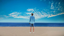 A person stands on a sandy beach looking at the ocean under a blue sky.