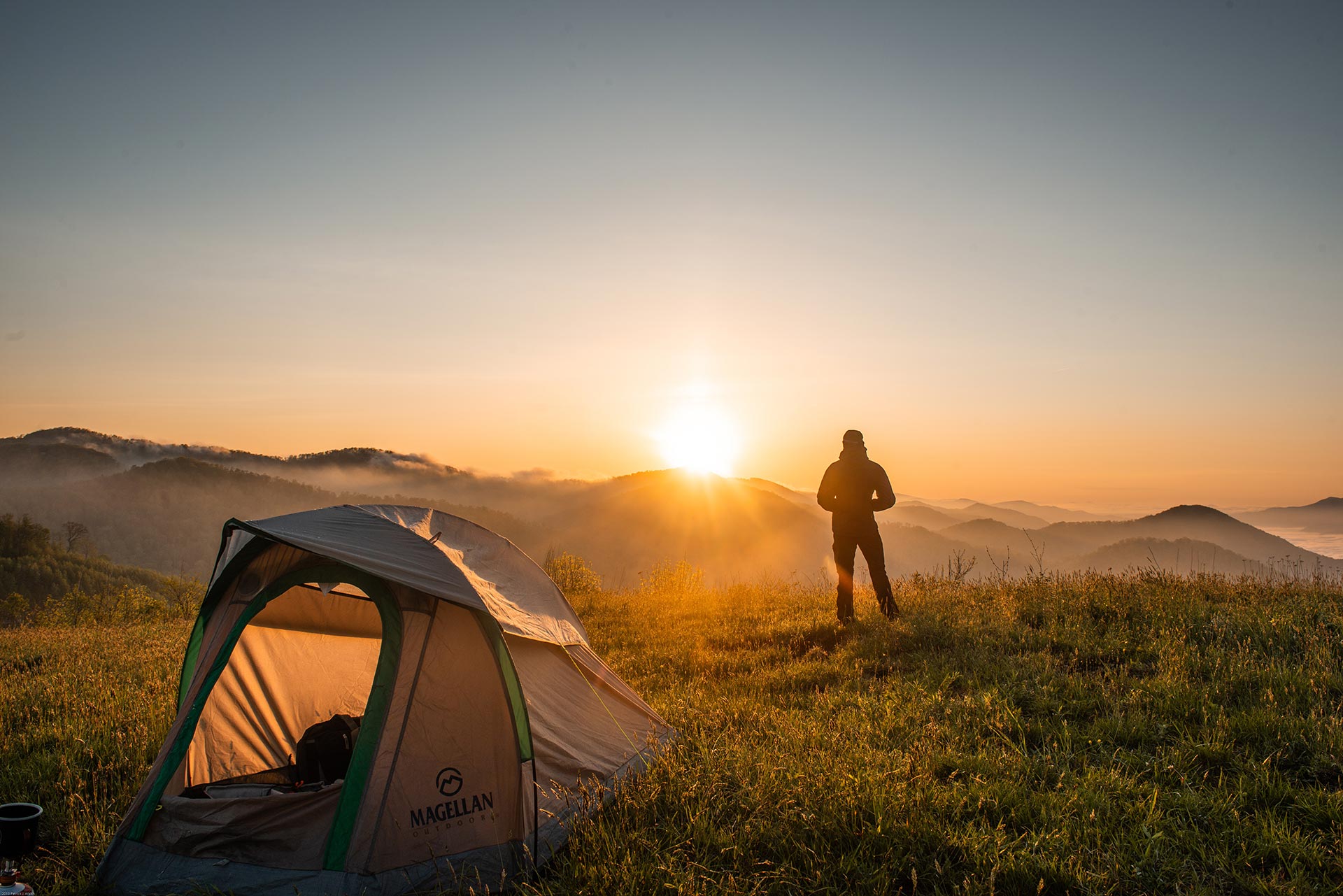 A person standing near a tent watching the sunrise over hills.